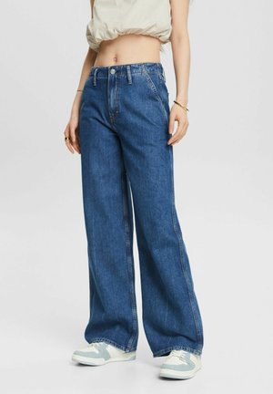 Person iført højtaljede, løstsiddende blå jeans, hvide og grå sneakers og en cropped beige top, der står mod en ensartet baggrund.