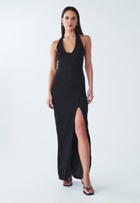 Svart maxiklänning med halterneck i texturerat tyg, sidospjäll, figursydd överdel och mjukt flödande kjol. Stylad med svarta sandaler med remmar.