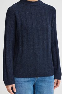 Maglione a maglia blu navy con collo alto, vestibilità ampia, texture a coste e maniche lunghe, abbinato a jeans azzurri.