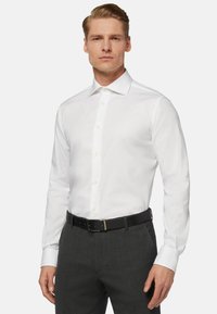 Camicia bianca a maniche lunghe con colletto classico, chiusura con bottoni sul davanti e una finitura in tessuto liscia, abbinata a pantaloni grigi scuri sartoriali e cintura nera.