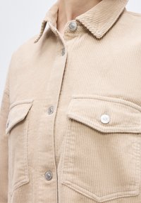 Μπεζ μπουφάν από κορντόρι με γιακά, κλιπ κουμπιά και δύο τσέπες στο στήθος με καπάκια, που φοριέται από ένα άτομο.