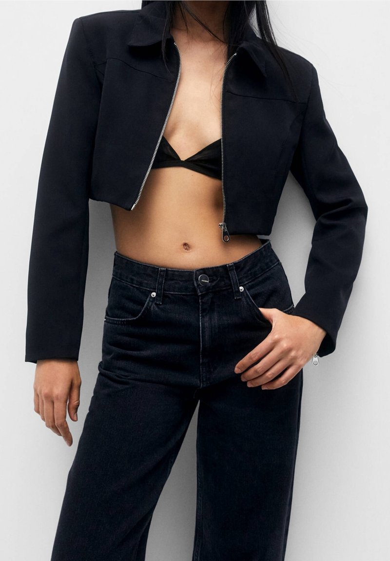 Giacca nera corta con zip frontale e colletto, abbinata a pantaloni in denim neri a vita alta. Il tessuto appare liscio e strutturato.