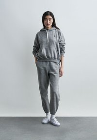 Νεαρή γυναίκα που στέκεται μέσα σε εσωτερικό χώρο, φοράει γκρι φούτερ με κουκούλα και ταιριαστό παντελόνι φόρμας, μαζί με λευκά αθλητικά παπούτσια, μπροστά σε απλή πρόσοψη τοίχου.