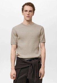 Beige gestrickter kurzärmeliger Pullover mit einem strukturierten Muster, Rundhalsausschnitt und geradem Schnitt, getragen mit dunklen Hosen und einem braunen Pullover, der um die Taille gebunden ist.
