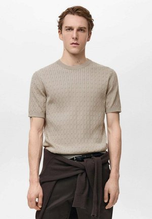 Beige gebreid kortemouwen sweater met een gestructureerd patroon, ronde hals en rechte pasvorm, gedragen met donkere broeken en een bruine sweater om de taille geknoopt.