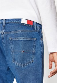 Sininen farkkutalje, jossa on takatasku ja joka on varustettu punaisella, valkoisella ja sinisellä bränditunnuksella, jossa on painettuna sana "JEANS".