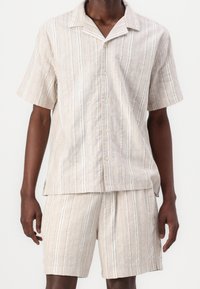 Camisa de manga corta de lino y pantalones cortos a juego en color beige claro con rayas verticales. Cuenta con cuello de solapa y cierre de botones. Tejido texturizado.