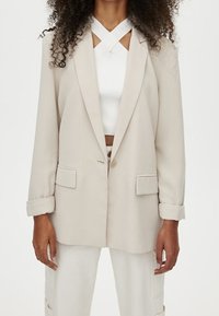 Blazer beige avec un seul bouton, revers à encoche, et deux poches avant. Porté sur un haut blanc avec un col croisé.