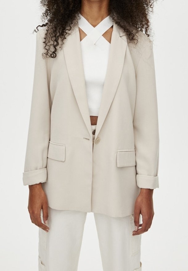 Blazer beige avec un seul bouton, revers à encoche, et deux poches avant. Porté sur un haut blanc avec un col croisé.