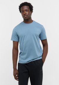 Jasnoniebieska koszulka z krótkim rękawem, wykonana z miękkiego materiału. Posiada okrągły dekolt i małe logo na klatce piersiowej. W połączeniu z ciemnymi spodniami.