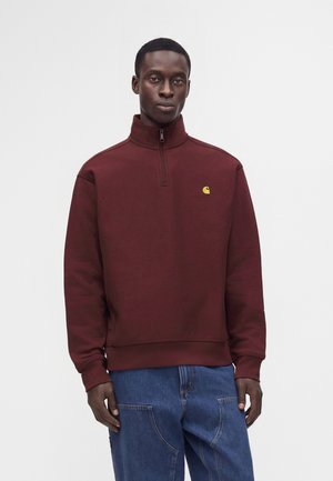Man draagt een bordeauxrood kwart-ritssweatshirt met een klein geel logo op de borst en blauwe jeans, staand tegen een effen witte achtergrond.