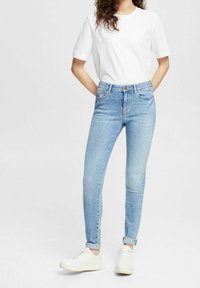 Vaqueros ajustados de color azul claro con una cintura alta, ligero desgastado, cinco bolsillos y puños enrollados, combinados con una camiseta blanca sin estampado y zapatillas blancas.