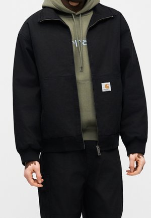 Homme portant une veste noire zippée par-dessus un sweat à capuche vert olive avec texte de logo, pantalon noir et zones de peau visibles sur les mains.