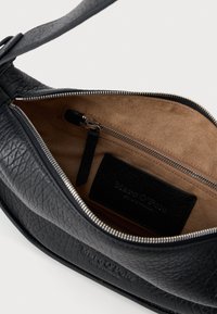 Sac en cuir noir à bandoulière avec une finition texturée. La fermeture éclair révèle un intérieur beige et une étiquette avec le nom de la marque.
