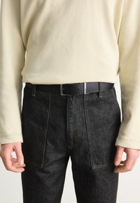 Cintura nera tessurizzata con fibbia argentata, indossata sopra pantaloni in denim scuro e una camicia a maniche lunghe color crema con una texture a coste.
