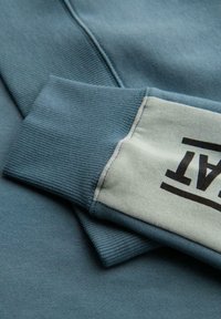 Detalle de sudadera de color azul claro con puños acanalados texturizados; un puño tiene una sección contrastante en gris claro con un logotipo negro audaz.