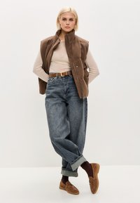 Chaleco marrón sin mangas sobre una blusa de manga larga beige, combinado con jeans de mezclilla claros de tiro alto y mocasines marrones.