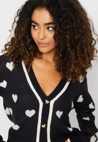 Zwarte cardigan met witte hartpatronen, met een V-hals, contrasterende afwerking en gouden hartvormige knopen. Zacht breimateriaal.