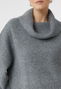 Femme portant un pull en tricot côtelé gris avec un col roulé large et replié, ainsi que de grandes boucles d'oreilles en or visibles.