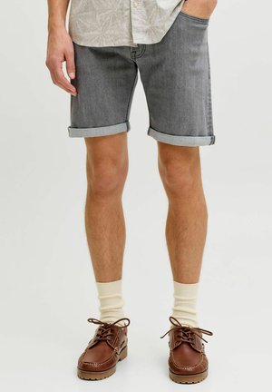 Man wearing grey rolled-up denim shorts, cream socks, and brown lace-up shoes, standing with one hand in pocket and the other relaxed.