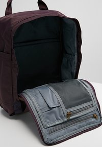 Ciemnofioletowy plecak z otwartą główną komorą i przednią klapą, która odsłania wiele zamykanych na zamek i siatkowych kieszeni organizacyjnych.