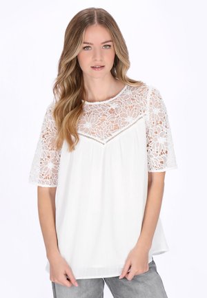 Femeie purtând o bluză albă cu detaliu floral din dantelă pe pieptul superior și pe mâneci, combinată cu pantaloni gri deschis, stând într-un cadru cu fundal alb.