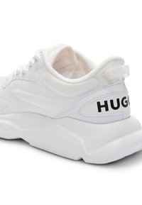 Valkoinen tennari verkkomateriaalilla ja kumipohjalla. Siinä on pehmustettu kaulus, musta "HUGO"-logo kantapäässä ja teksturoitu sivuosa paremman pidon takaamiseksi.