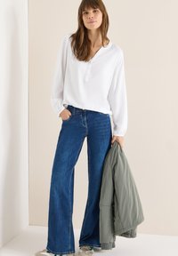 Chemise blanche à manches longues avec un col boutonné, accompagnée d'un jeans évasé bleu foncé. Une veste vert clair est tenue dans une main.