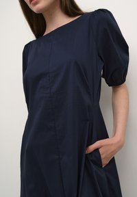Mörkblå klänning gjord av slät tyg, med fyrkantig halsringning, puffade korta ärmar och sidofickor. Enkel, elegant design.