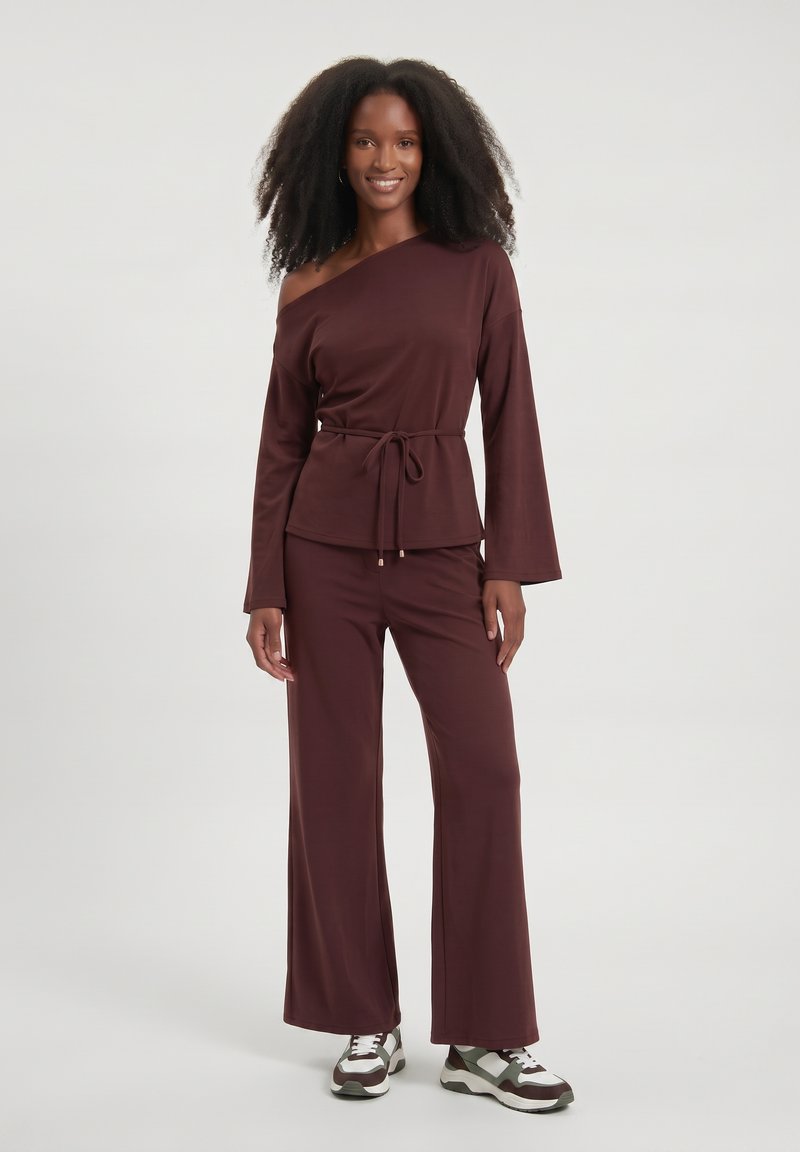 Femme aux cheveux bouclés naturels souriant, portant un haut bordeaux à une épaule avec ceinture à la taille, un pantalon large et des baskets blanches sur un fond uni.
