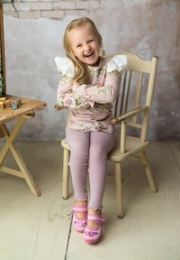 Kind in einem floral gemusterten, pinken Langarmoberteil mit Spitzenakzenten, trug pinke Leggings und pinke Schuhe mit Herzdetails und sitzt auf einem Holzstuhl.