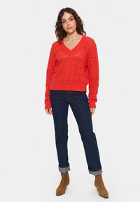 Pull rosso lavorato a maglia con scollo a V e motivo traforato, abbinato a jeans scuri arrotolati ai polsini e stivaletti alla caviglia in suede marroni.