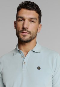 Lichtblauwe polo gemaakt van katoen, met een kraag, drie knopen en een hexagonaal logo op de linkerkant van de borst. Glad textuur.