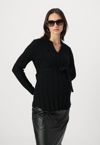 Svart ribbestrikket genser med V-hals, belted i livet, kombinert med en skinnende svart imitert skinnskjørt. Modell som bruker oversized svarte solbriller.