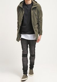 Giacca verde oliva con cappuccio, indossata sopra una maglietta nera e una maglietta bianca sotto. Jeans skinny grigio scuro con dettagli a coste e scarpe slip-on grigie.