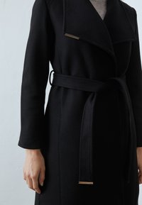 Cappotto di lana nero con ampi revers, vita con cintura e un accento dorato sul colletto. Presenta maniche lunghe e una texture liscia.