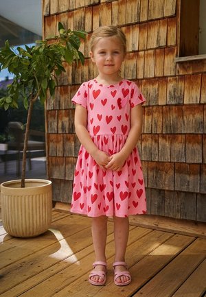 Ein junges Mädchen steht auf einem Holzdeck, trägt ein rosa Kleid mit roten Herzen und passende Sandalen neben einer Topfpflanze.