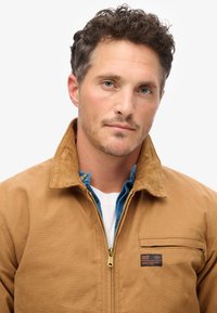 Bruine corduroy jas met een hoge kraag, ritssluiting aan de voorkant en borstzak; gedragen over een blauwe spijkerhemd en een wit T-shirt.