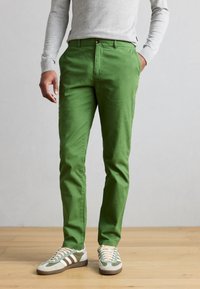 Πράσινο βαμβακερό παντελόνι από τουίλ με στενή εφαρμογή, που διαθέτει μπροστινό κούμπωμα με κουμπιά και πλαϊνές τσέπες, συνδυασμένο με γκρι αθλητικά παπούτσια.