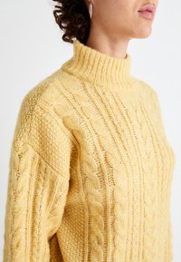 Pull en laine jaune pâle à maille torsadée avec un col montant, présentant un motif texturé et une silhouette ample.