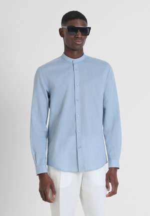 Camicia azzurra chiara a maniche lunghe con colletto arrotondato, logo sul petto e tessuto morbido. Indossata con occhiali da sole neri oversize.