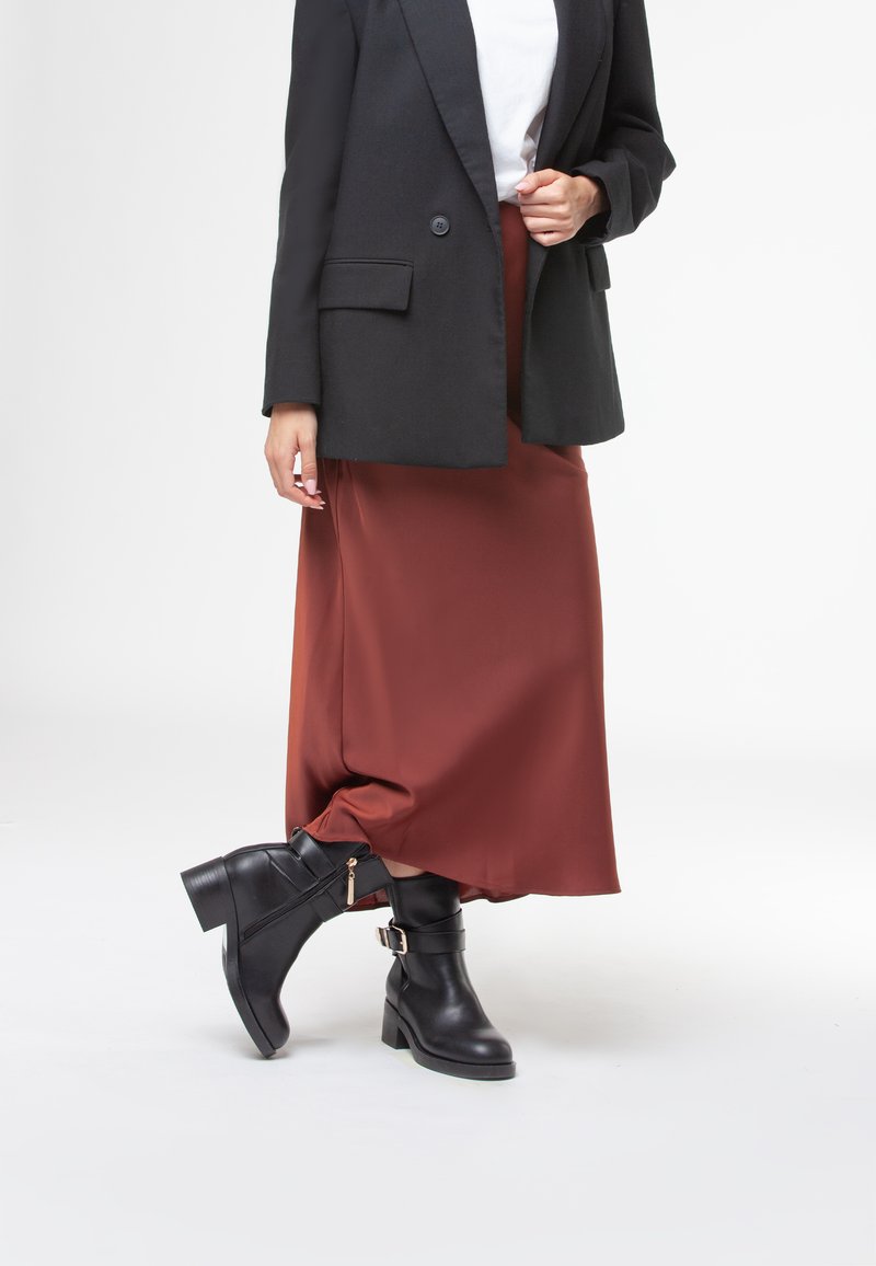 Stivali neri in pelle fino alla caviglia con tacco a blocco, cinturino con fibbia e cerniera laterale, abbinati a una lunga gonna in satin bordeaux e a un blazer nero oversize.
