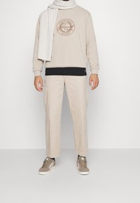 Béžová mikina s kulatým logem, černé tričko s krátkým rukávem, béžové kalhoty a šedé tenisky. Bílý šátek volně visí kolem krku, má jemnou texturu.