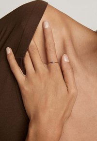 Anillo de oro con una pequeña gema azul, llevado en una mano que descansa sobre una prenda marrón, destacando la simplicidad y elegancia del anillo.