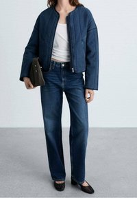 Veste en denim bleu au design court, associée à un jean bleu taille haute. Le mannequin tient un sac à main foncé et porte des ballerines noires.