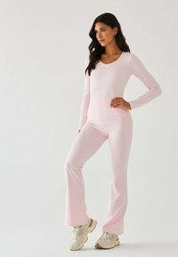 Bladoróżowa bluzka z długim rękawem i dopasowane rozszerzane spodnie wykonane z elastycznego materiału. Luźny krój, gładka faktura. Noszone z beżowymi butami sportowymi.