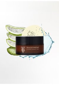 Ronde bruine pot met een zwart deksel met het etiket "True Botanicals At-Home Facialist Peel." Op de achtergrond groene aloëverabladeren en een waterplons.