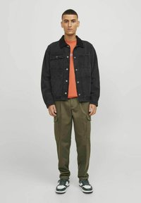 Schwarze Denimjacke mit Fronttaschen, oranges T-Shirt, olivgrüne Cargo-Hose und dunkle Sneakers. Modell steht vor einem schlichten Hintergrund.