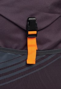 Paarse stoffen rugzak met een gestructureerd oppervlak, voorzien van een zwarte kunststof speld en een oranje bevestigingsriem. Subtiel grijs patroon langs de onderkant.