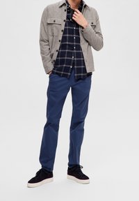 Šedá košile s knoflíky přes námořnicky modrou kostkovanou košili, doplněná námořnicky modrými kalhotami a tmavými teniskami. Klíčový důraz na textury a barvy.